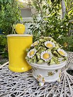Photo of yellow ceramic canister with yellow apple lid next to covered ceramic daisy bowl on outdoor white table.