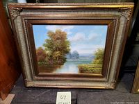 Framed oil painting showing a river scene with trees and a partly cloudy sky.