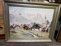 Framed signed oil painting showing a mountain village with houses, church steeple, mountains in background.
