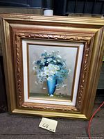 Full view of the framed floral still life painting showing white and blue flowers in a blue vase with an ornate gold frame.