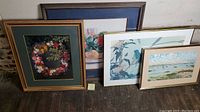 Four framed prints depicting floral wreath, still life flowers, bird scene, and coastal boat scene arranged on wooden floor.