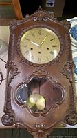 Front view of antique Reguladora wall clock showing carved wooden case, round dial with Roman numerals, clock hands and pendulum visible through glass.