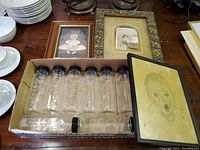 Box containing nine Evenflo clear glass baby bottles with black lids, one bottle lying on its side. Three framed baby-related artworks visible behind the box.