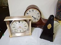Set showing antique Seth Thomas wooden mantle clock, Bulova battery desk clock with dedication plaque, and vintage mechanical metronome.