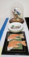 Porcelain Blue Jay figurine under glass dome, stack of four duck-themed dishes, and Asian fish themed artwork displayed on a white surface.