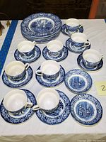 Photo showing multiple cups and saucers, plates stacked on a table in blue and white transferware Liberty Blue pattern.