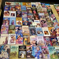 A photo showing about 59 vintage paperback romance and pulp fiction books arranged on a flat surface. Covers are visible, showcasing colorful illustrated art typical of 1970s and 1980s genre novels.