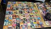 Wide shot showing approximately 80 vintage paperback romance novels arranged on a flat surface. The books have colorful illustrated covers and appear well-used.