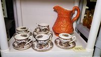 Photo showing five transferware teacups with matching saucers and an orange ceramic pitcher on shelf.