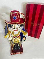 Front view of the hand painted musketeer nutcracker with detailed clothing and accessories, standing next to the original box.