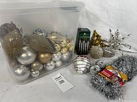 Photo showing plastic bin filled with assorted Christmas ball ornaments, box of icicle ornaments, large unique ornaments, and silver tinsel garland.