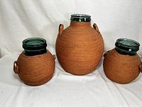 Set of 3 VIRESA green glass demijohn bottles with woven brown wrap and wooden bangle handles, against white fabric background.