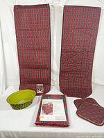Two red and green plaid woven cotton table runners standing vertically, one olive green woven basket with liner, one clear glass tumbler, one red and green plaid pot holder on flat surface, and one unopened plaid tablecloth package laid flat.
