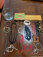 Assorted beaded jewelry including coral necklaces and bracelets, pearl, jade, shell beaded bracelets, a cribbage board with playing cards, and a clear glass bowl arranged on a wooden surface.