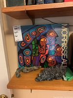 Book 'Morrisseau' cover with colorful indigenous art and two rough metal fragments on wooden shelves.