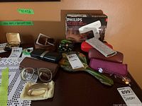 Full lot showing Philips hair dryer in box, various eyeglasses and cases, hand mirror, trinket box, leather belt, and silver bracelet on lace tablecloth.
