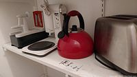 Shows white apple peeler, mixer parts, KitchenAid red kettle, and Chef stainless steel toaster arranged on a shelf.