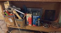 Wide view of workbench lower shelf with various boxes, cords, tools, power bars, miscellaneous plumbing fittings and Mastercraft utility pump in box plus propane torch tank in box.