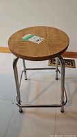 Metal stool with round wood veneer top and four chrome-finished legs, taken on tiled floor near wall.