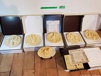 Six cream-colored relief decorative wall plates displayed together with original boxes and one on a display stand; all approximately 8.5 inches in diameter.