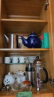 Full cabinet shelf view showing all items together: stainless steel teapot, dark blue ceramic teapot, boxed items (possibly tea), porcelain espresso cups and saucers, glass and black French press, white jar with holes, glass sugar container, and stainless steel thermal flask.