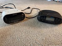 Photo showing two alarm clocks; a white Sony AM/FM digital clock radio and a black Timex digital alarm clock on carpet.
