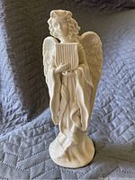 White ceramic angel statue standing upright on a rounded base, holding a lyre, detailed wings and flowing robes visible.