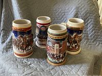 Four Budweiser holiday themed ceramic beer steins arranged on a cloth background. Each stein shows horse-drawn carriage winter scenes with colorful detailing.