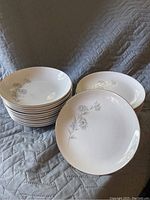 Stack of 11 white salad plates with a subtle floral design on a quilted grey fabric background