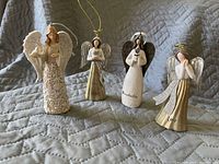 Four Christmas angel figurines displayed on a quilted fabric background. All have white and gold tones, with distinct wings and inscriptions on the front.