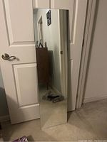 Full length rectangular mirror leaning against a white door, showing clear reflection of room interior including door handle, floor, and wall decorations.