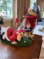 Photo of three golf-themed Santa Claus figurines in Christmas attire positioned on a table near a window, showing one putting, one holding a flag, and one swinging a golf club