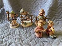 Five small vintage Hummel porcelain figurines depicting children engaged in various activities, sitting, playing instruments, and carrying sticks.