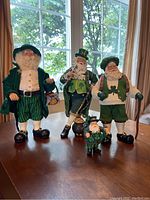 Four Irish Santa Claus figurines on table near window. Each dressed in green themed attire with white beards and various accessories including baskets, coins and cane.