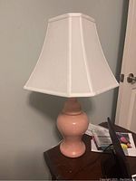Photo showing a pink ceramic table lamp with a white fabric shade placed on a wooden surface near a door.