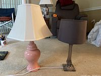 Photo shows two table lamps on a carpeted floor. One with a pink ceramic base and cream square lampshade, the other with a transparent base and dark brown cylindrical lampshade.