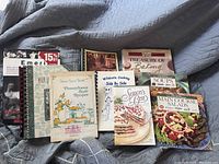 Photo of a collection of 9 recipe and cooking books with various styles and titles, placed on a gray-blue quilted fabric background.