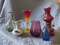 Six assorted vintage glass pieces on gray quilted fabric background including vases, pitchers, and an oil lamp.