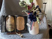 Image showing a decorative open book display with a painted scenic image and poem alongside a pot holding silk flowers with various colors and an American flag ribbon attached.
