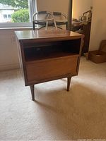 Front view of wooden nightstand showing drawer and open shelf, positioned on carpeted floor near window and walker.