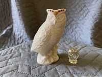 Photo showing two owl figurines on a gray quilted surface, one larger white ceramic with red accents and one smaller clear crystal with green eyes.