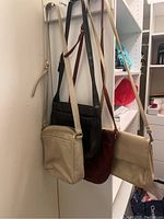Photo showing four purses hanging in a closet. Beige, black, burgundy, and cream colored purses with straps visible.