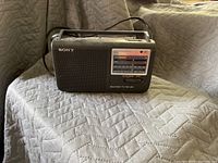 Photo of a black Sony portable radio showing front speaker grill, frequency dial and tuning display with AM/FM bands, placed on a quilted surface