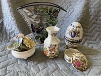 Photo of various decorative glass and porcelain items including butterfly-themed glass vase, porcelain flower bouquet, porcelain vase, trinket box, and decorative egg container.