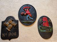 Three vintage cast iron fire insurance plaques, one depicting a fireman in red cape, another with four clasped hands and number, and a third with red fire hydrant and letters 'F A'.