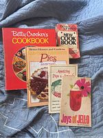 Five vintage cookbooks laid flat on a blue quilted fabric backdrop showing front covers. Titles visible include Betty Crocker's Cookbook, Better Homes and Gardens Pies, New Cook Book, an appetizer dips and spreads booklet, and a Joys of Jell-O dessert booklet. Covers are worn and edges frayed.