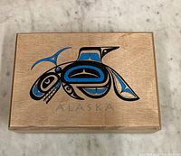 Top view of wooden box showing Alaska native art design with black and blue stylized bird motif and the word ALASKA.