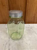 Front view of hexagonal green tinted glass jar with silver metal screw lid on a marble surface against wooden backdrop.