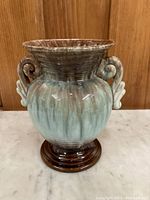 Front view of ceramic vase showing bulbous body, two curled handles, and base with striped gradient glaze in brown, green, and blue tones.