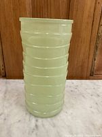 Front view of the opaque pale green ribbed glass vase on a white surface in front of wood paneling.